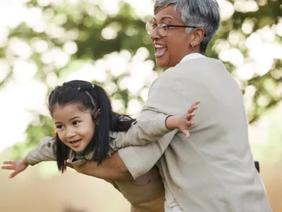 A grandmother joyfully playing with her granddaughter, lifting her in the air like an airplane in an outdoor setting.