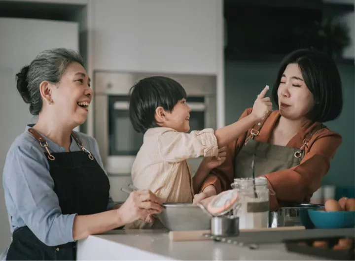 A young boy playfully touches his mother's nose with flour while cooking together in the kitchen with his grandmother.